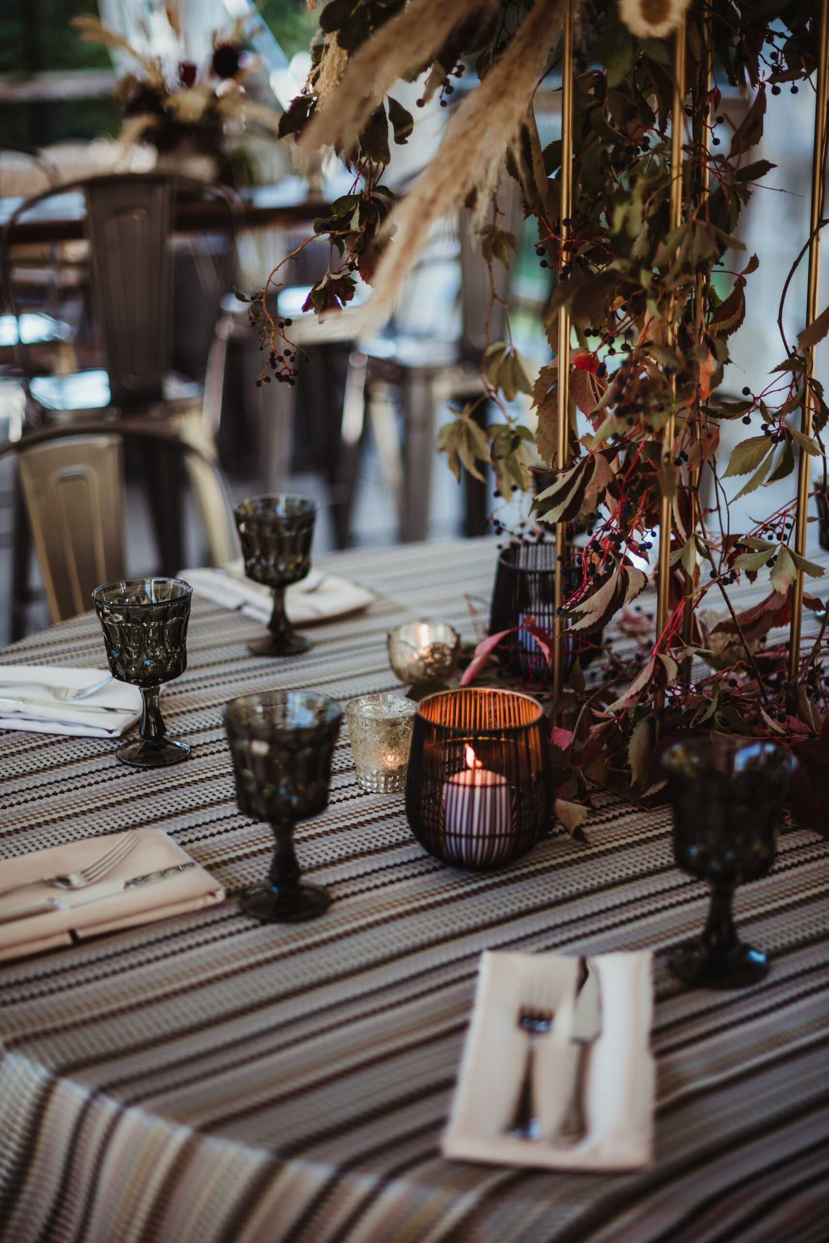 Dark glasses and napkins with silverware are set around a moody fall table setting with candles and leaves.